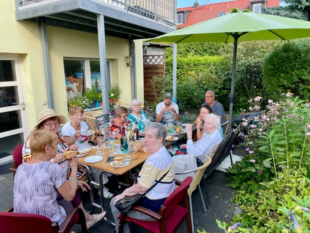 Sommerfest im Garten: rund zehn Bewohnerinnen und Bewohner sitzen an einer langen Tafel unter einem grünen Sonnenschirm, umgeben von Beeten und Sträuchern.