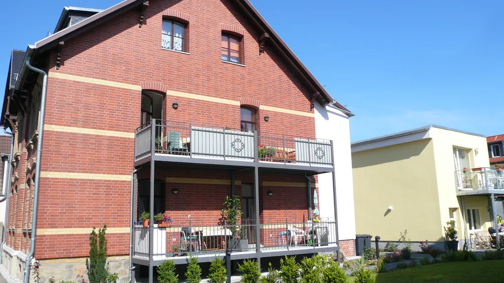 Blick vom Hof auf das Seniorenhaus: ein zweigeschossiger Klinkerbau mit hellen Zierbändern, davor Balkone und überdachte Terrassen mit Sitzgruppen, ringsum frisch bepflanzte Beete und ein gepflasterter Weg.